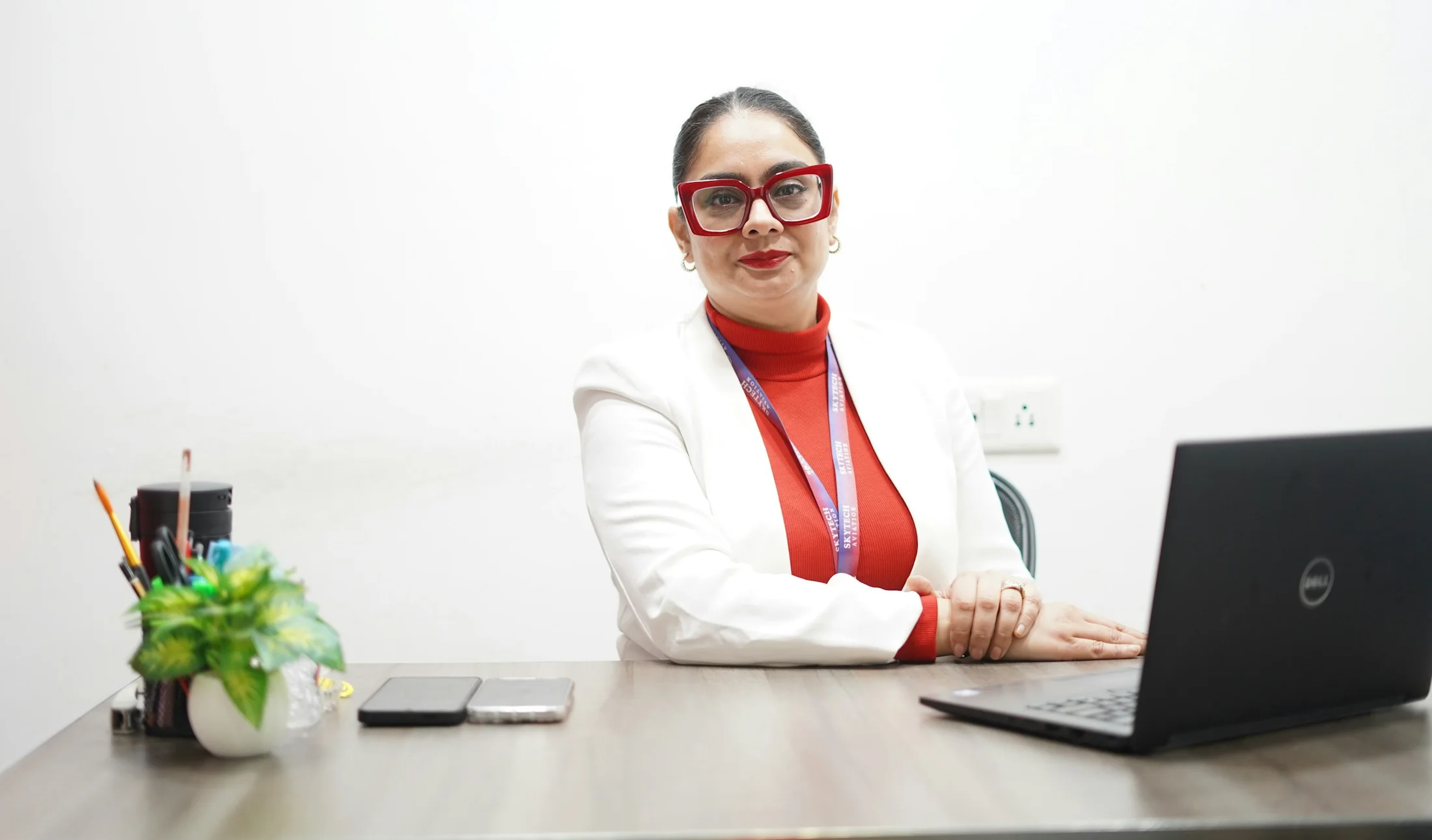 An Indian woman in red glasses and a white blazer, seated at a desk with a laptop.